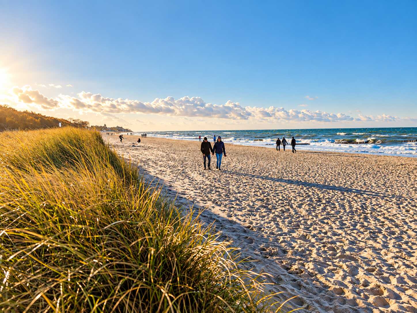 Ein sonniger Ostseestrand mit goldenen Dünen, sanften Wellen und Spaziergängern, die die ruhige Küstenatmosphäre genießen.
